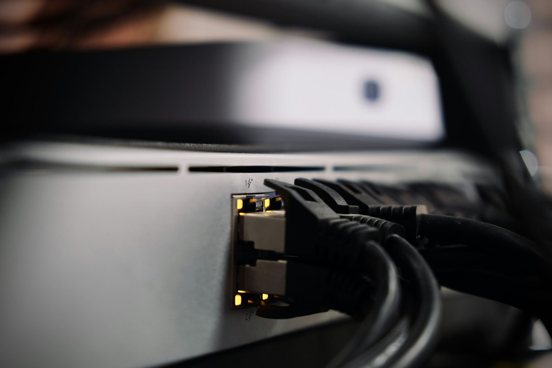 Black network cables plugged into the ports of a silver network switch, with the port lights glowing and the background softly out of focus